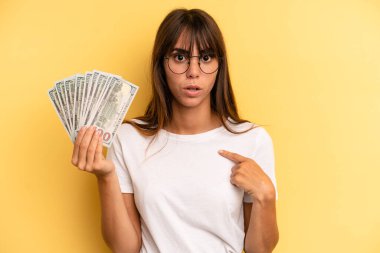hispanic woman looking shocked and surprised with mouth wide open, pointing to self. dollar banknotes concept