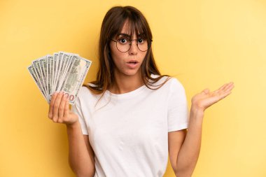hispanic woman looking surprised and shocked, with jaw dropped holding an object. dollar banknotes concept