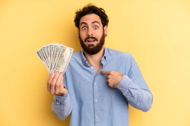 Şok olmuş ve ağzı açık, kendini işaret eden genç bir adam. Dolar banknotları konsepti