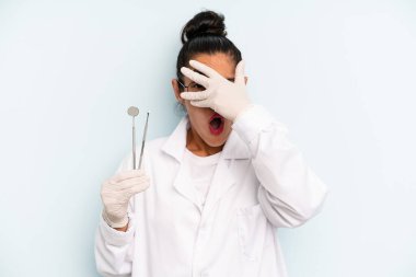 hispanic woman looking shocked, scared or terrified, covering face with hand. dentist concept
