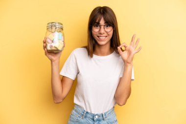 hispanic woman feeling happy, showing approval with okay gesture. candies bottle concept
