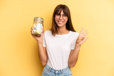 hispanic woman smiling and looking friendly, showing number four. candies bottle concept