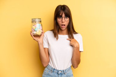 hispanic woman looking shocked and surprised with mouth wide open, pointing to self. candies bottle concept