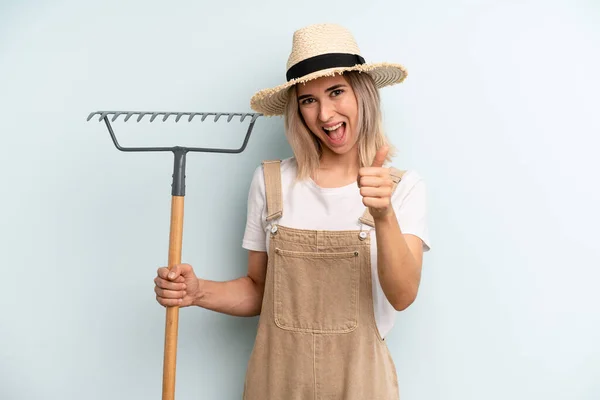 blonde woman feeling proud,smiling positively with thumbs up. farmer and rake cocnept