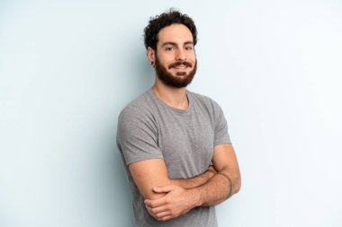 Genç, sakallı bir adam kameraya çapraz kollarıyla gülümsüyor ve mutlu, kendinden emin, tatmin olmuş bir yüz ifadesi var.