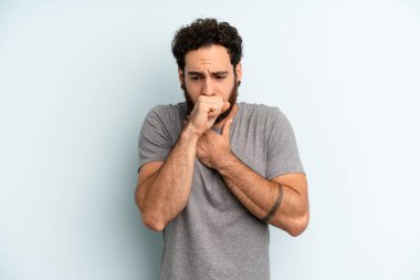 Genç, sakallı, boğazı ağrıyan ve grip belirtileri gösteren ağzı kapalı öksüren bir adam.