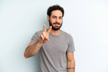 Genç, sakallı adam gülümsüyor ve arkadaş canlısı görünüyor. İki ya da iki numarayı gösteriyor. Elleri önde, geriye doğru sayıyor.