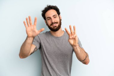 Sakallı genç adam gülümsüyor ve arkadaş canlısı görünüyor. 8 ya da 8 numarayı gösteriyor. Elleri önde, geriye doğru sayıyor.