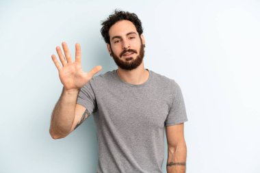 Genç, sakallı bir adam gülümsüyor ve arkadaş canlısı görünüyor. 5 ya da 5 numarayı gösteriyor. Eli önde, geriye doğru sayıyor.