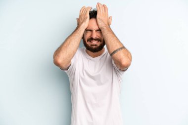 Genç, sakallı bir adam stresli, endişeli, depresif ve hayal kırıklığına uğramış hissediyor, iki elini de başa kaldırıyor.