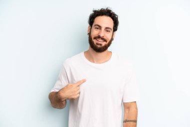 Genç yetişkin sakallı adam gururlu, kendinden emin ve mutlu görünüyor, gülümsüyor ve kendini işaret ediyor ya da bir numaralı işareti yapıyor.