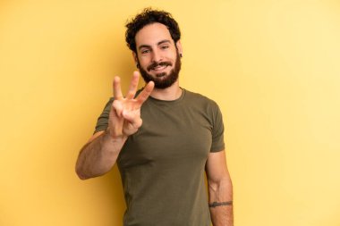 Genç, sakallı bir adam gülümsüyor ve arkadaş canlısı görünüyor. Elleri önde ve geriye doğru sayıyor.