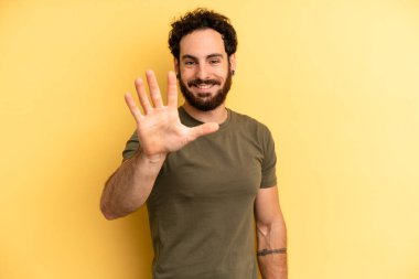 Genç, sakallı bir adam gülümsüyor ve arkadaş canlısı görünüyor. 5 ya da 5 numarayı gösteriyor. Eli önde, geriye doğru sayıyor.