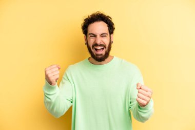 Genç, sakallı, mutlu, şaşırmış ve gururlu bir adam. Büyük bir gülümsemeyle başarıyı bağırıyor ve kutluyor.