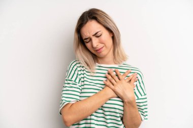 young adult blonde woman looking sad, hurt and heartbroken, holding both hands close to heart, crying and feeling depressed