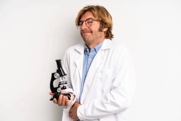 blond adult man shrugging, feeling confused and uncertain. scientist with a microscope