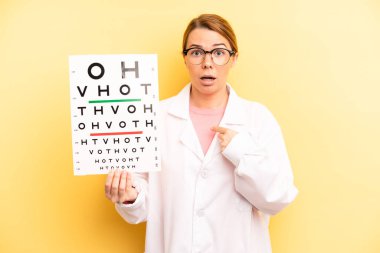 pretty blonde young woman looking shocked and surprised with mouth wide open, pointing to self. optical vision test concept