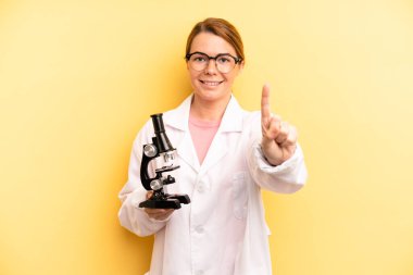 pretty blonde young woman smiling and looking friendly, showing number one. scients student with a microscope concept