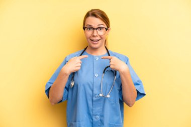 pretty blonde young woman feeling happy and pointing to self with an excited. physician or doctor student concept