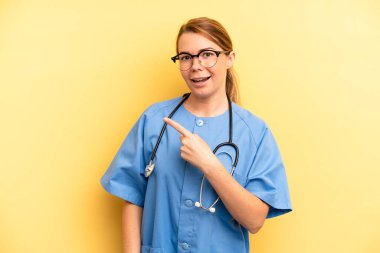 pretty blonde young woman looking excited and surprised pointing to the side. physician or doctor student concept
