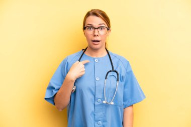 pretty blonde young woman looking shocked and surprised with mouth wide open, pointing to self. physician or doctor student concept