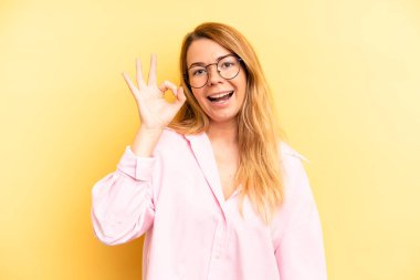 Sarışın, güzel bir kadın kendini başarılı ve tatmin olmuş hissediyor, ağzı açık gülümsüyor, elle işaret veriyor.