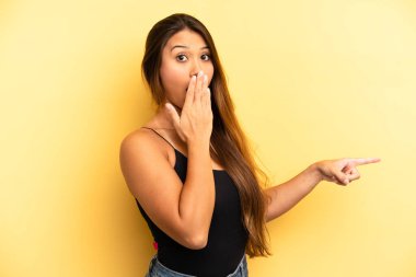 Asyalı güzel kadın kendini mutlu, şaşkın ve şaşırmış hissediyor. Ağzını eliyle kapatıyor ve yanal fotokopi alanını işaret ediyor.