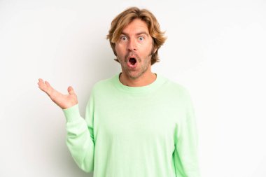 Sarışın, yetişkin bir adam şaşırmış ve şok olmuş görünüyor, çenesi düşmüş, elinde açık bir elle bir nesne tutuyor.