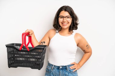 İspanyol güzel kız mutlu bir şekilde gülümsüyor, eli kalçasında ve kendinden emin. boş alışveriş sepeti kavramı