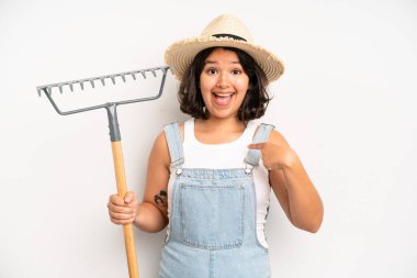 hispanic pretty girl looking shocked, scared or terrified, covering face with hand. farmer and rake concept