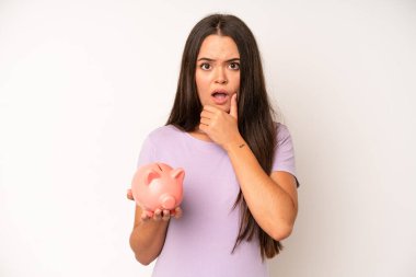 hispanic pretty woma smiling happily, waving hand, welcoming and greeting you. piggy bank savings concept