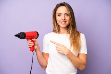 Güzel, beyaz bir kadın, neşeli bir şekilde gülümsüyor, mutlu hissediyor ve bir tarafı işaret ediyor. saç kurutma makinesi konsepti
