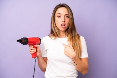 Ağzı açık, kendini işaret eden oldukça beyaz bir kadın şok olmuş ve şaşırmış görünüyor. saç kurutma makinesi konsepti