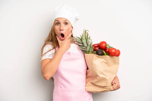 pretty caucasian woman with mouth and eyes wide open and hand on chin. chef with a market vegetables bag