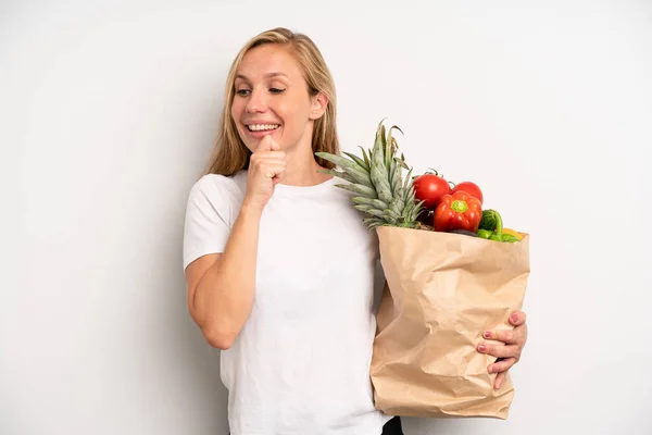 pretty caucasian woman smiling with a happy, confident expression with hand on chin. chef with a market vegetables bag