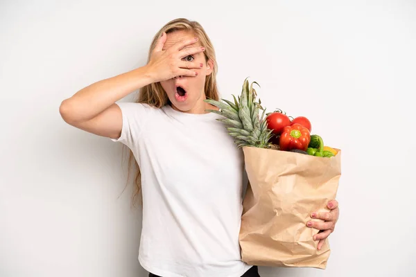 pretty caucasian woman looking shocked, scared or terrified, covering face with hand. chef with a market vegetables bag
