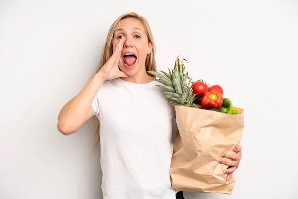 pretty caucasian woman feeling happy,giving a big shout out with hands next to mouth. chef with a market vegetables bag