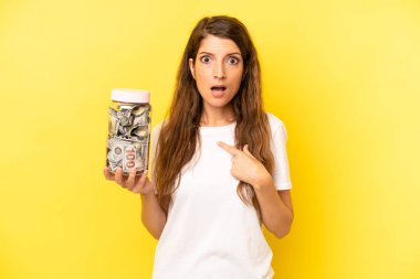 pretty caucasian woman looking shocked and surprised with mouth wide open, pointing to self. dollar banknotes savings
