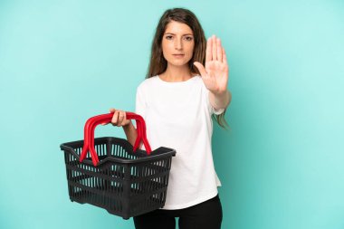 pretty caucasian woman looking serious showing open palm making stop gesture. empty market basket concept