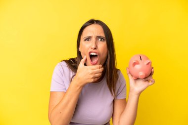 Ağzı ve gözleri açık ve eli çenesinde olan Latin kökenli genç bir kadın. domuz kumbarası tasarruf kavramı