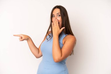 İspanyol güzel kadın kendini mutlu, şaşkın ve şaşırmış hissediyor. Ağzını eliyle kapatıyor ve yanal kopya alanını işaret ediyor.