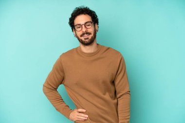 Genç sakallı adam mutlu bir şekilde gülümsüyor, eli kalçasında ve kendinden emin, olumlu, gururlu ve arkadaş canlısı.