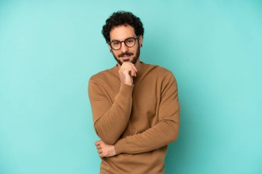 Genç sakallı adam mutlu görünüyor ve gülümsüyor. Elleri çenesinde, merak ediyor ya da soru soruyor. Seçenekleri karşılaştırıyor.