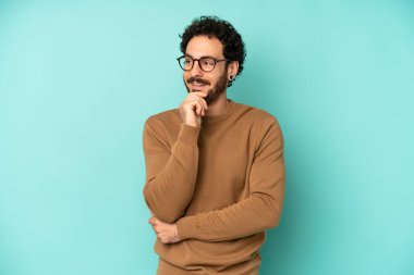 Genç sakallı bir adam mutlu, kendinden emin bir ifadeyle gülümsüyor. Elleri çenesinde, merak ediyor ve yan tarafa bakıyor.