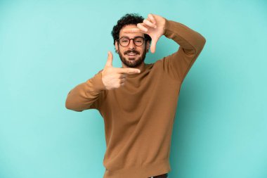 Genç sakallı adam mutlu, arkadaş canlısı ve pozitif hissediyor, gülümsüyor ve elleriyle bir portre veya fotoğraf çerçevesi yapıyor.