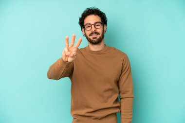 Genç sakallı adam gülümsüyor ve arkadaş canlısı görünüyor. Üç ya da üç numarayı gösteriyor. Elleri önde, geriye doğru sayıyor.