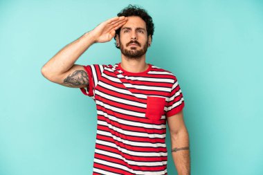 Genç sakallı bir adam saygı göstererek, onur ve vatanseverlik adına askeri bir selamlama ile kamerayı selamlıyor.