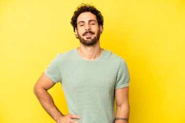 Genç sakallı adam mutlu bir şekilde gülümsüyor, eli kalçasında ve kendinden emin, olumlu, gururlu ve arkadaş canlısı.
