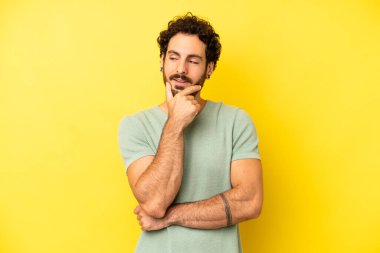 Genç sakallı bir adam mutlu, kendinden emin bir ifadeyle gülümsüyor. Elleri çenesinde, merak ediyor ve yan tarafa bakıyor.