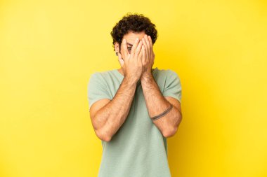 Genç sakallı adam elleriyle yüzünü kapatıyor, parmaklarının arasından şaşkın bir ifadeyle yan tarafa bakıyor.